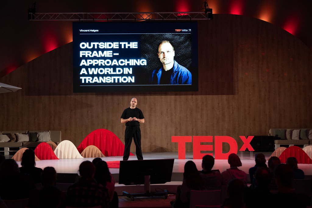 A speaker on stage in front of the slide “Outside the frame: approaching a world in transition”