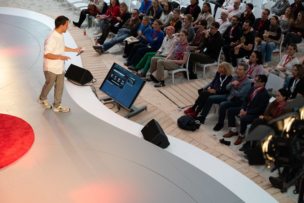 A speaker in a white shirt on the TEDxMitte stage in front of the audience